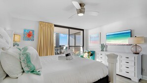 Habitación de hotel con cama, ventilador de techo, ventana con vistas a edificios, televisor y cómoda.