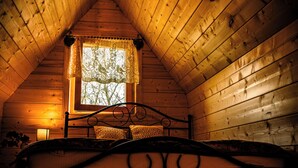 A cozy attic bedroom with a wooden sloped ceiling, a single bed with a metal frame, a small window with a lace curtain, and a hanging light fixture.
