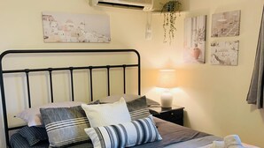 A neatly made bed with a variety of pillows and a blanket, a bedside lamp, and wall art. - Airport Cottage House (Spata-Artemida)