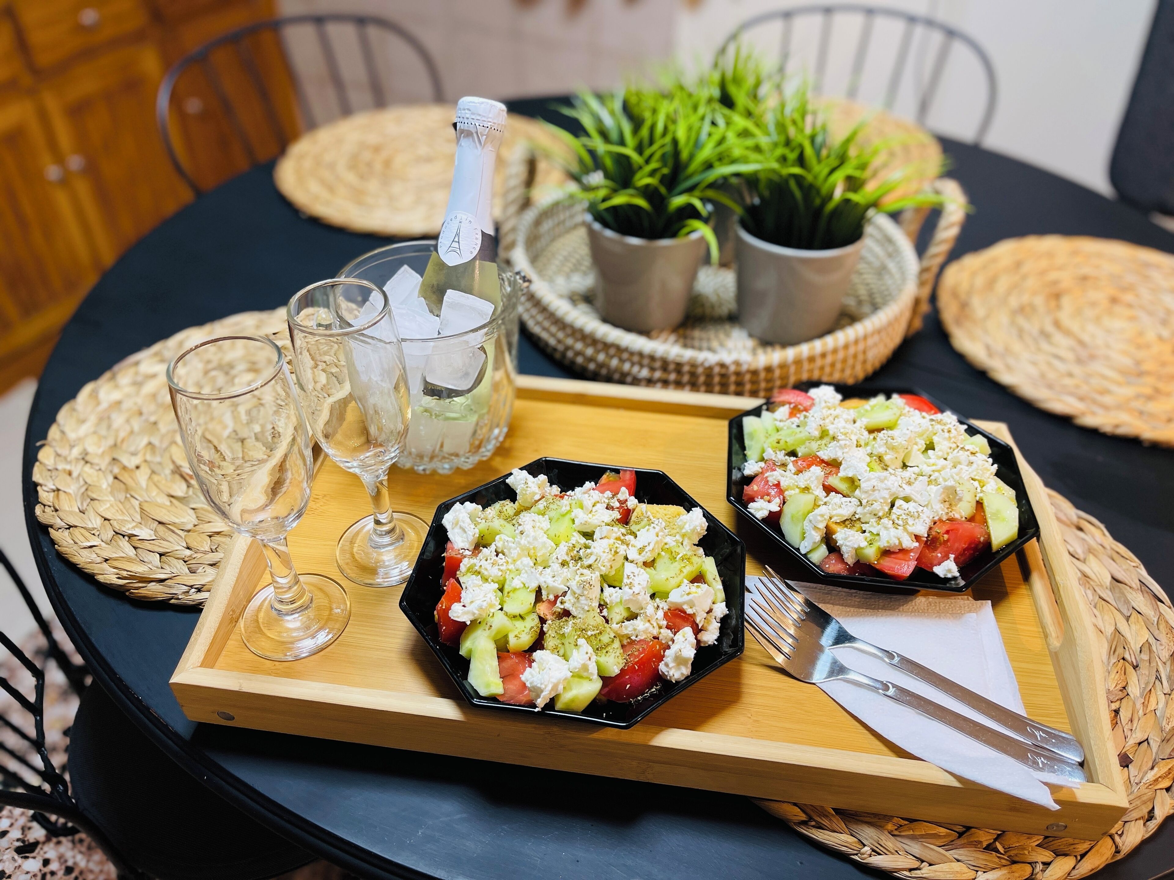 A round dining table with two black plates of salad, a bottle of champagne, and two glasses.