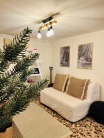 Una sala de estar acogedora con un sofá blanco, una mesa de centro y un árbol de Navidad. Hay dos cuadros en la pared y un ventilador de techo con luces.