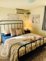 Una cama bien hecha con una manta beige mullida, una colcha azul y varias almohadas. Hay una lámpara de noche, una planta pequeña y una cesta con objetos en la mesita de noche.