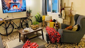A cozy living room with a television displaying a fireplace scene, a bicycle, a coffee table with snacks, a grey armchair, and a window with curtains. - Airport Cottage House (Spata-Artemida)