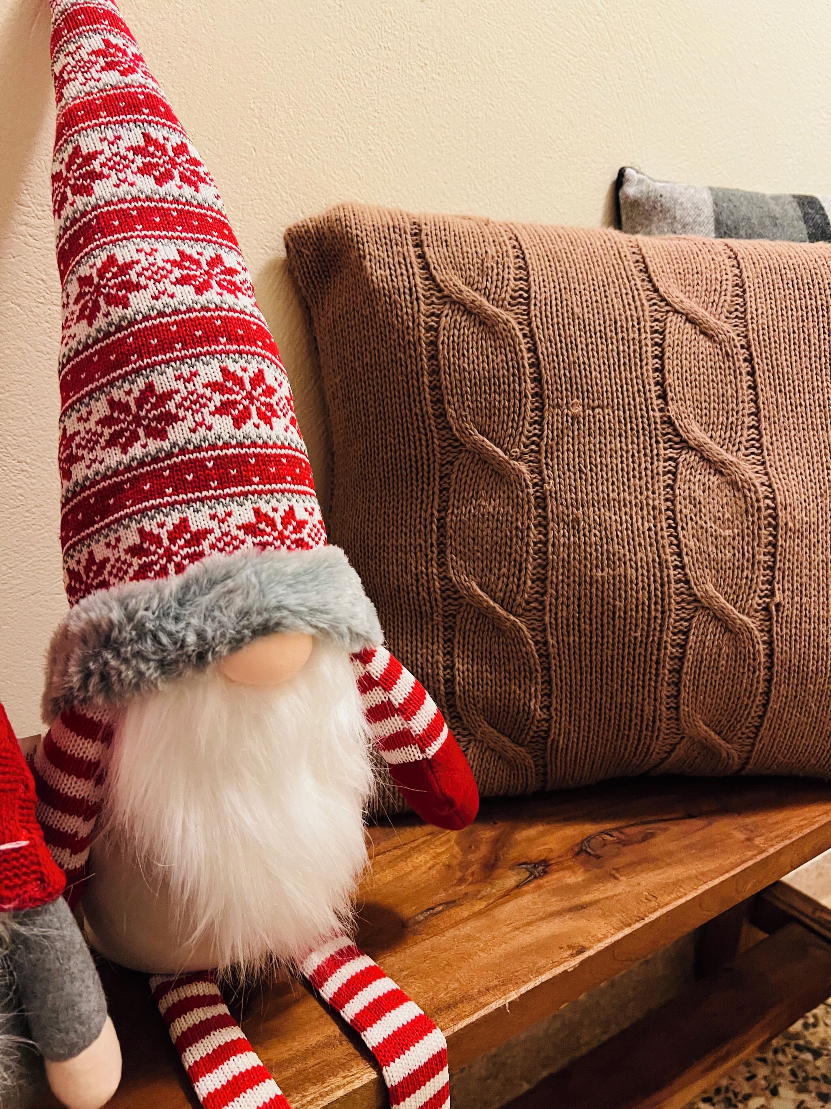 A gnome doll with a red and white striped hat and scarf, placed on a wooden bench next to a brown knitted pillow.