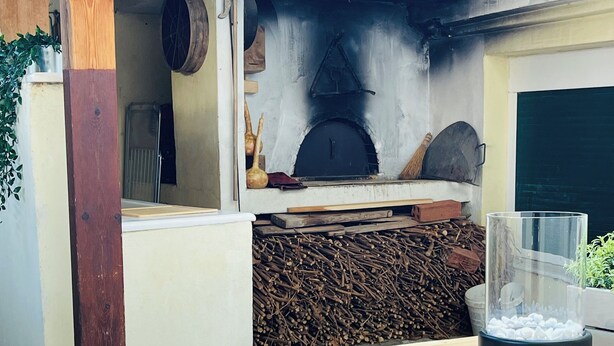 A wooden table with woven placemats, a glass container, and a rustic outdoor setting with a wood-burning stove. - Airport Cottage House (Spata-Artemida)