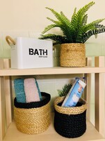 Un estante de baño con un recipiente blanco etiquetado “BATH KEEP CLEAN”, una cesta tejida con una toalla y una planta en maceta.
