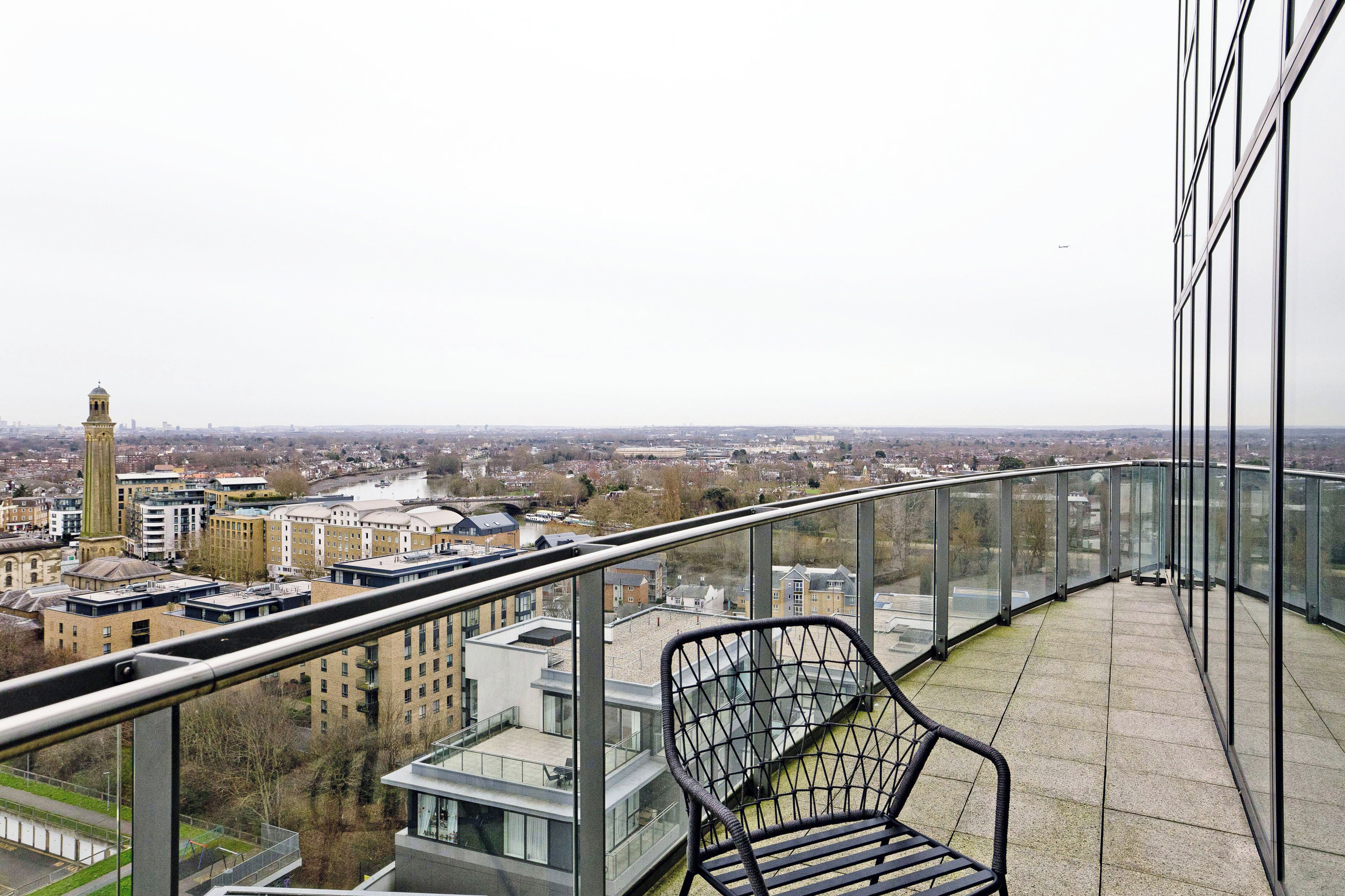 Un balcón con barandilla de vidrio que ofrece vistas a un paisaje urbano, con edificios modernos y una torre.