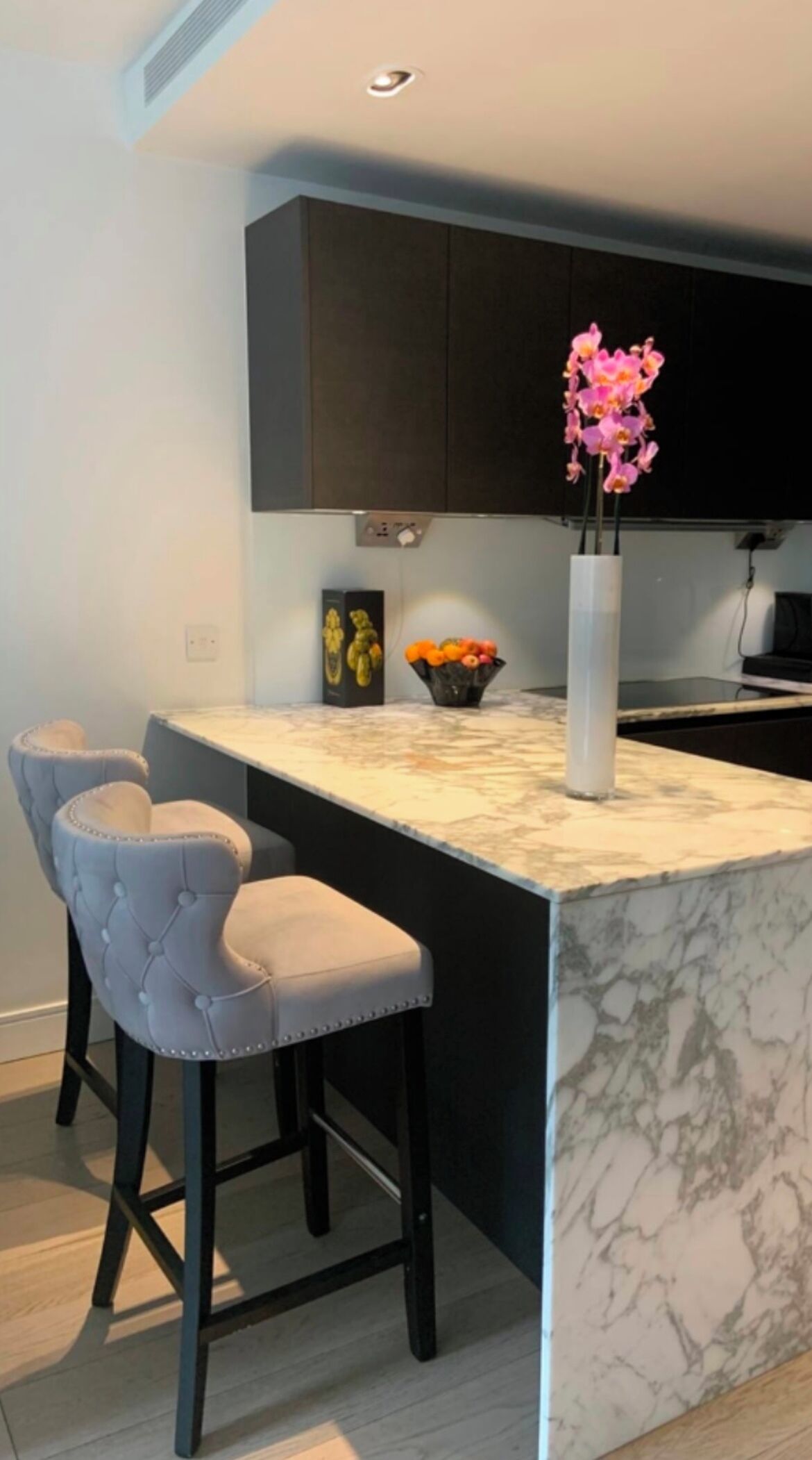 A modern kitchen with a marble island, two bar stools, dark cabinets, and a vase with flowers.