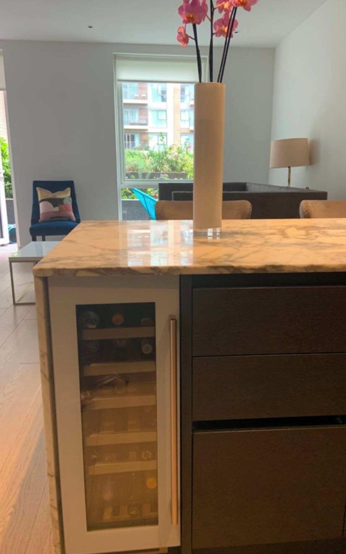 Une cuisine dotée d’un plan de travail en marbre, d’un réfrigérateur à vin intégré et offrant une vue sur un balcon aménagé avec du mobilier d’extérieur.