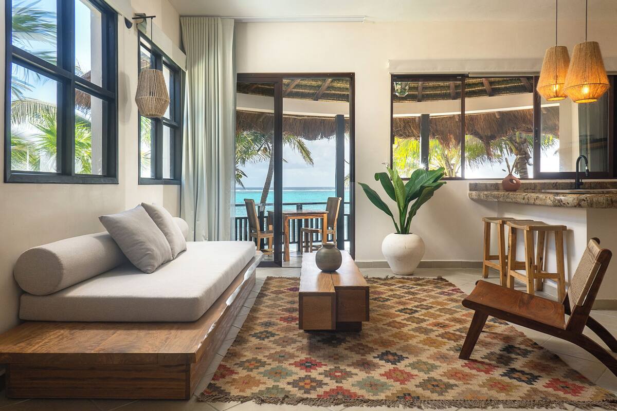 A modern living room with a sofa, a coffee table, and a bench. There is a large window with a view of the ocean and palm trees.