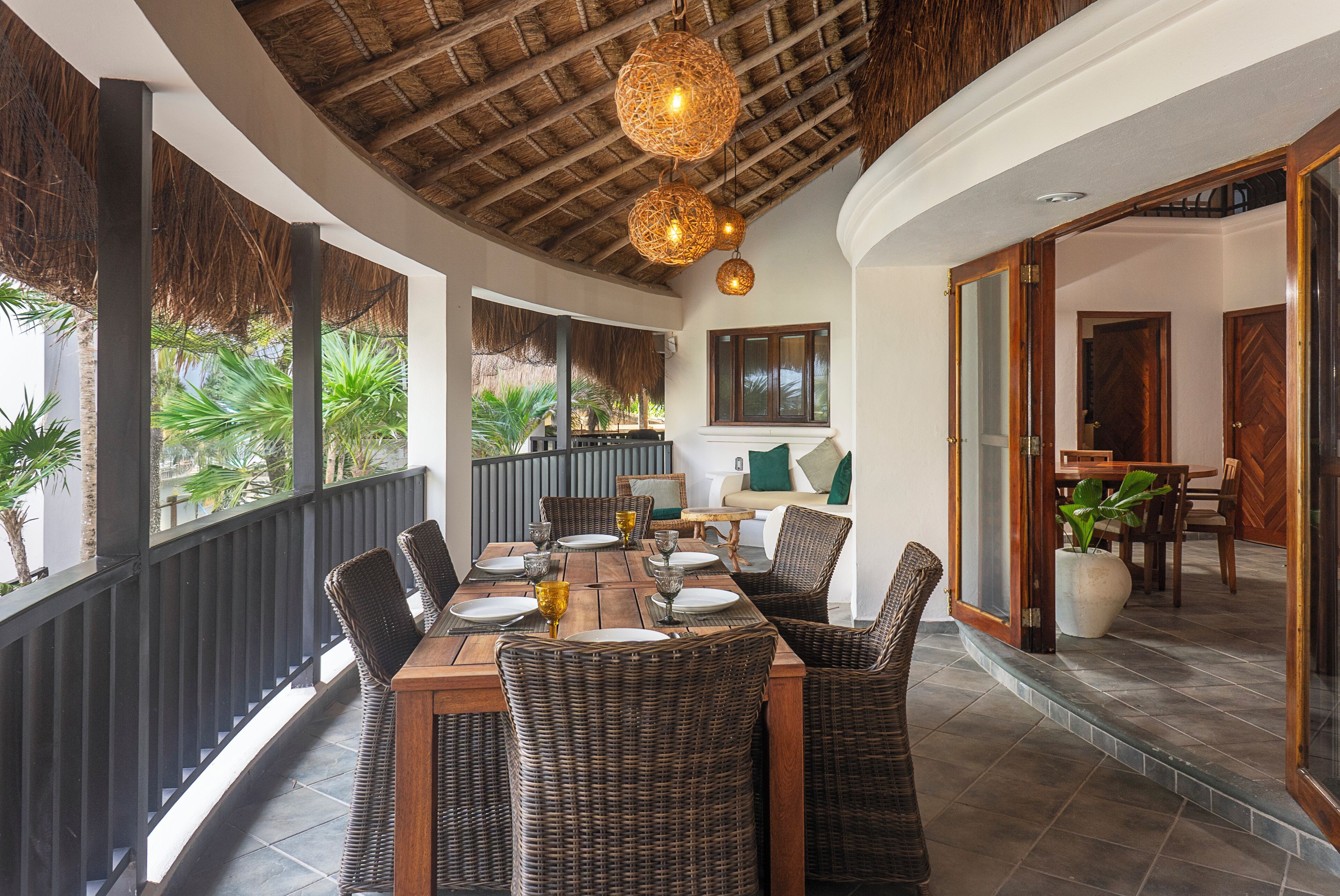 A dining area with a wooden table and wicker chairs, a ceiling with a thatched design, and a view of a garden through large windows.