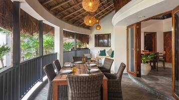 Un comedor con mesa de madera y sillas de mimbre, techo con diseño de paja y vistas a un jardín a través de amplios ventanales.