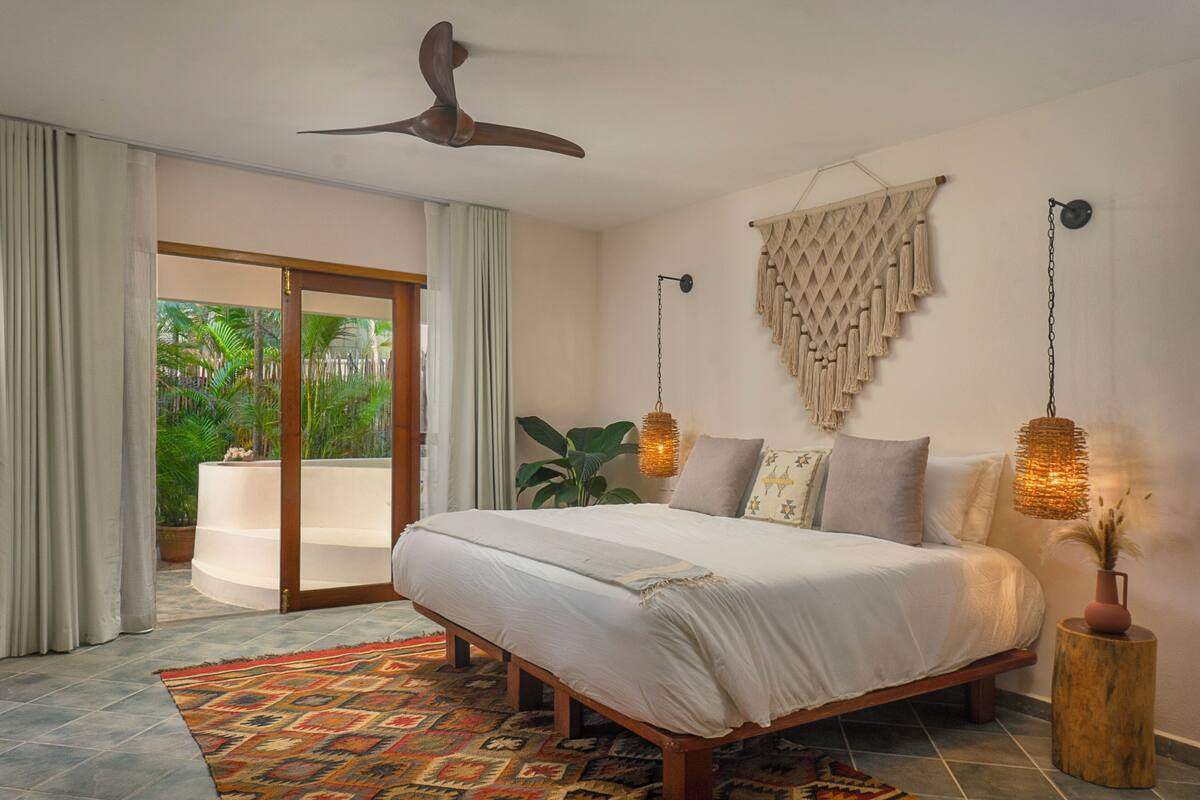 A bedroom with a bed, a ceiling fan, a large window, and a view of greenery outside.