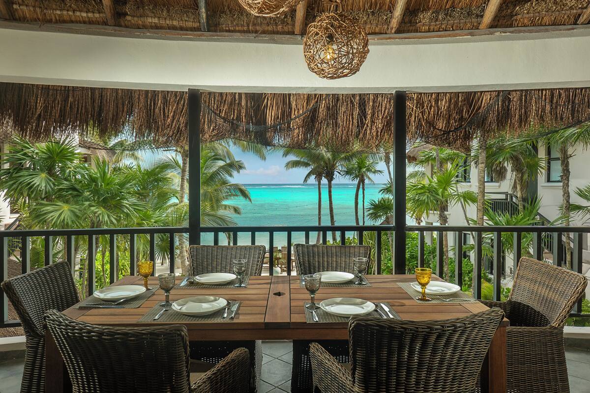 A dining area with a table set for four, overlooking a beach with palm trees and turquoise water.