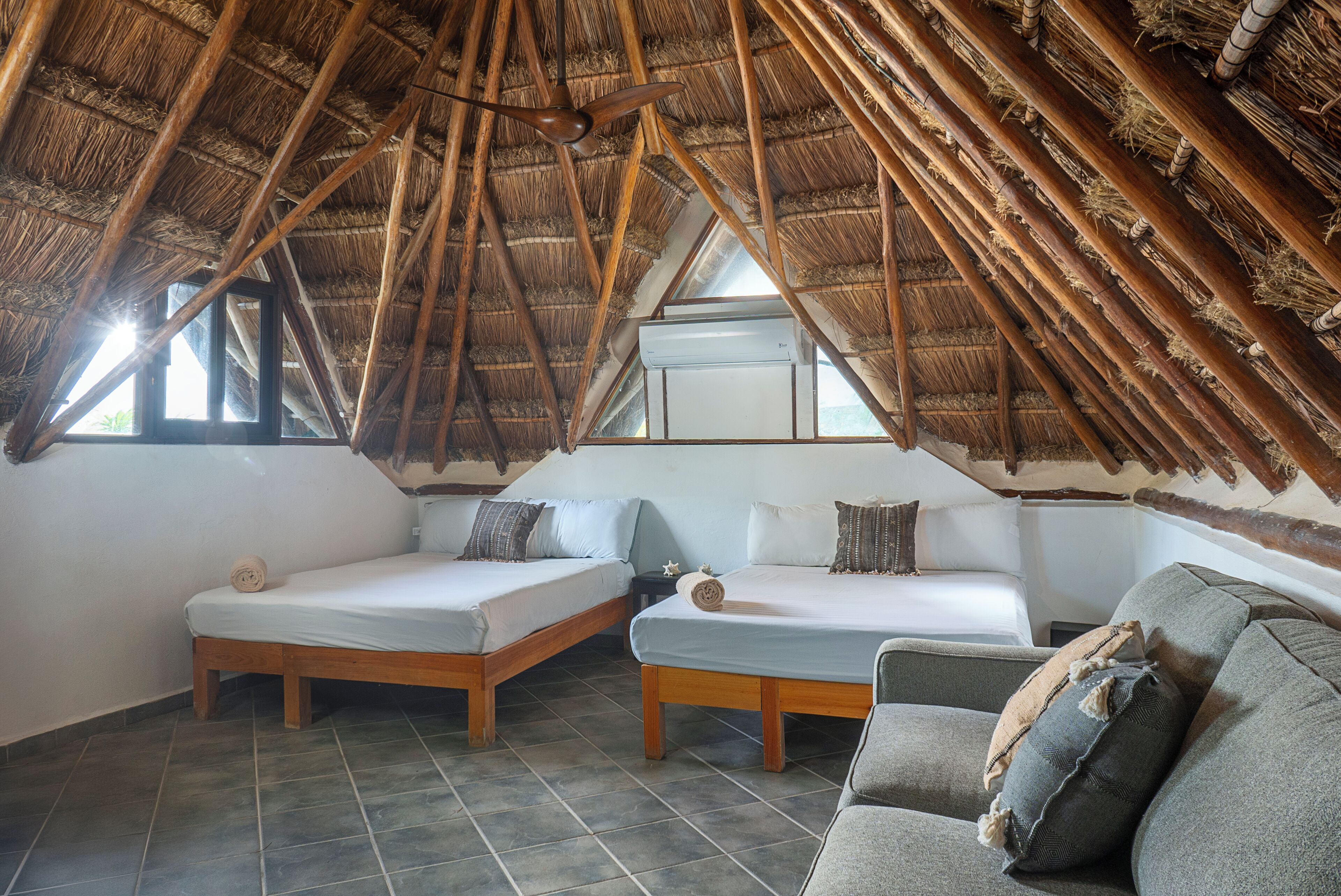 A cozy attic bedroom with a thatched roof, two wooden beds, a grey sofa, and a window providing natural light.