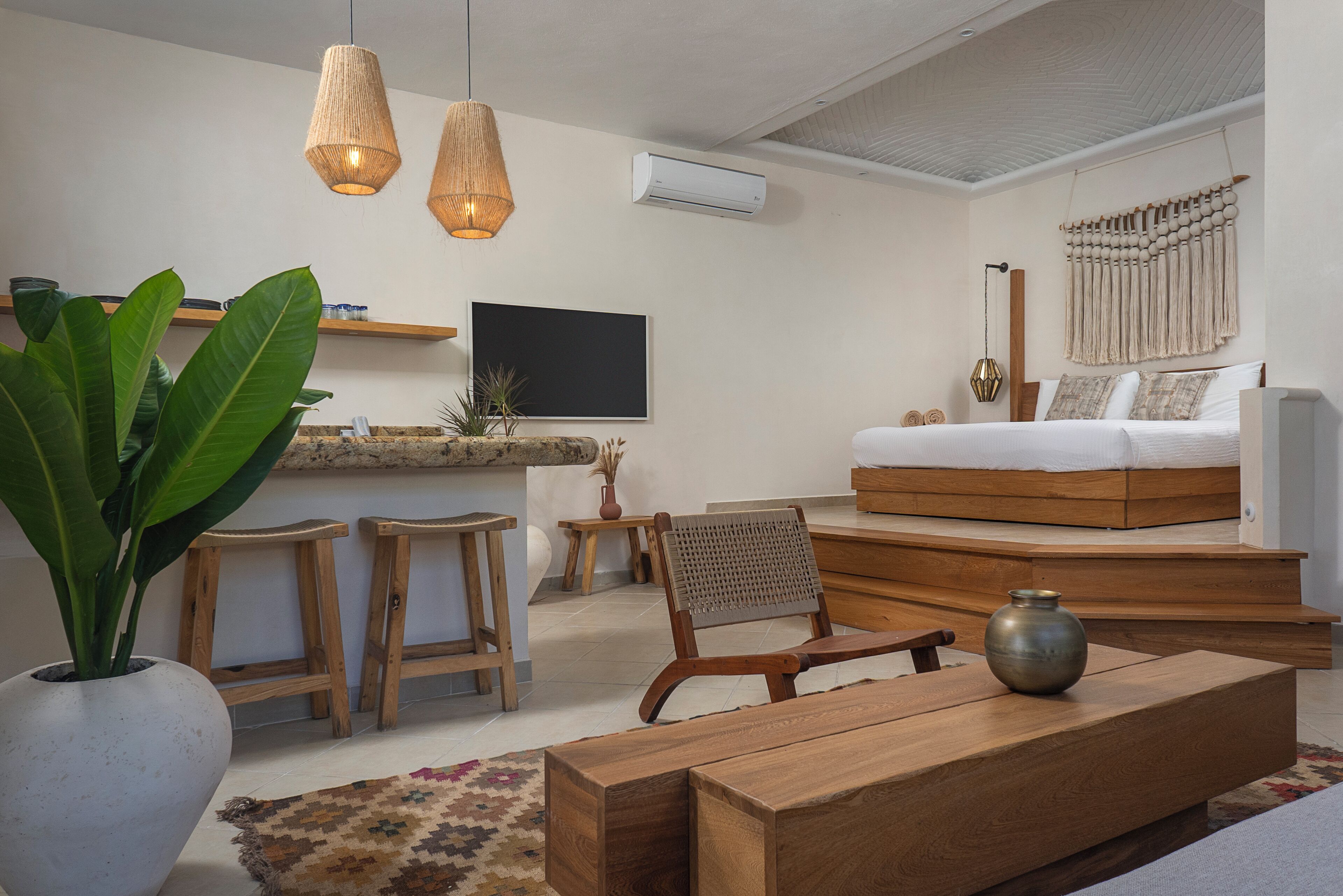 A modern living room with a wooden sofa, a coffee table, and a TV mounted on the wall. There are two wooden stools and a large potted plant.