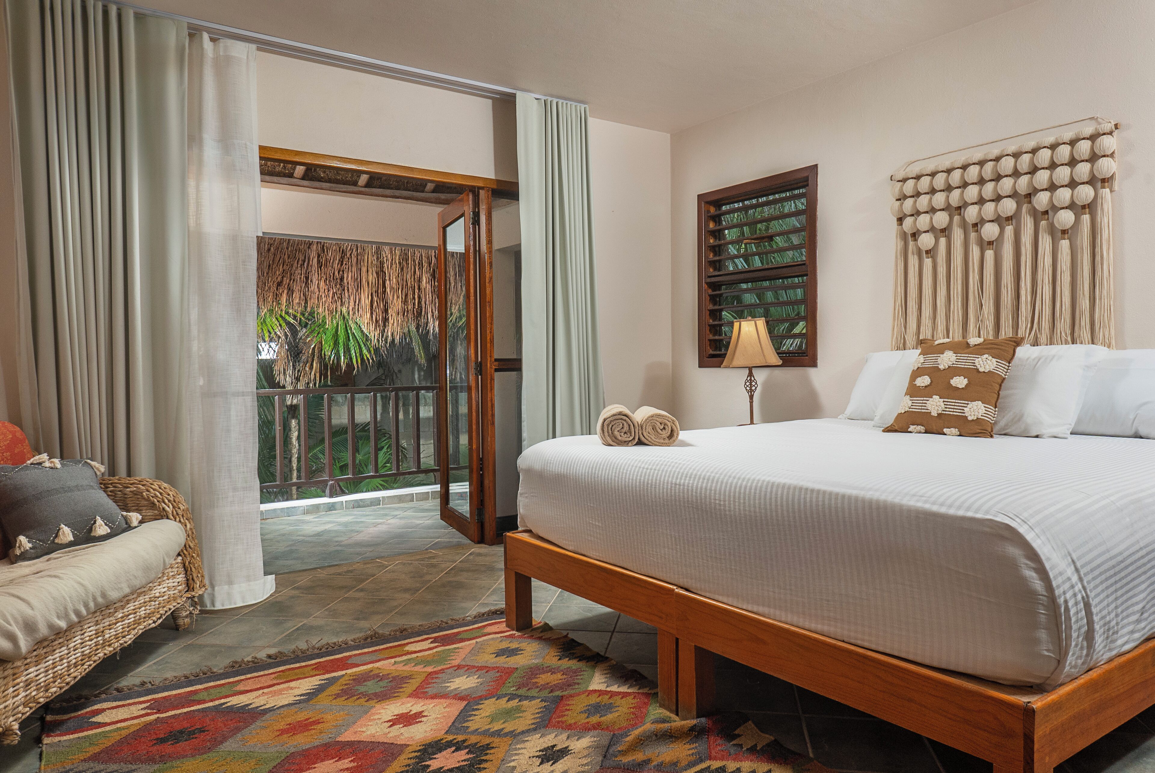Un dormitorio con cama, sofá, alfombra estampada, ventana con cortinas, lámpara de pared y un cuadro enmarcado.
