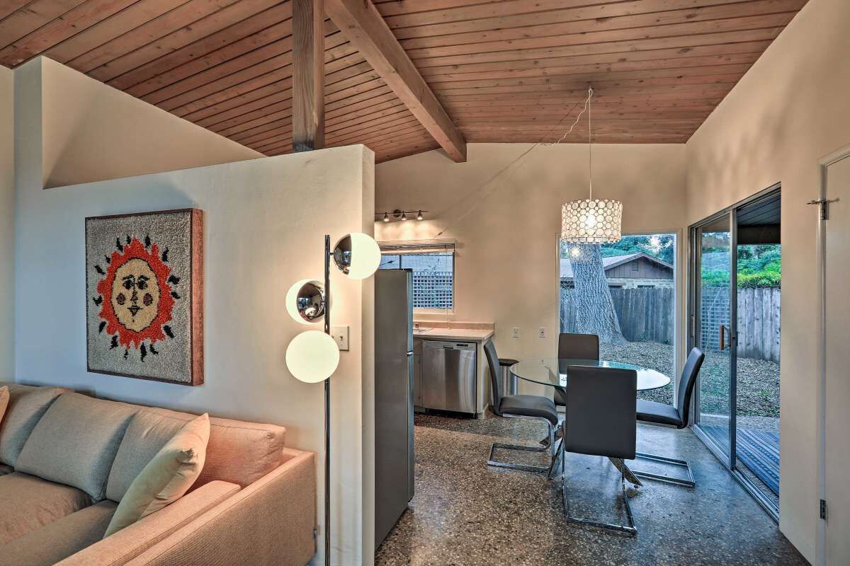 Un intérieur moderne avec un plafond en bois, un coin repas avec une table et des chaises en verre, une cuisine équipée d’appareils électroménagers en acier inoxydable et une porte-fenêtre coulissante donnant sur un jardin.