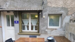 Un edificio rústico de piedra con una mesa y sillas de madera, una ventana pequeña con cortinas y un cartel que indica el número 61.