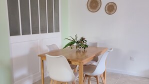 Un comedor con mesa de madera, sillas blancas, una planta en maceta, una lámpara de techo tejida y decoración en la pared.