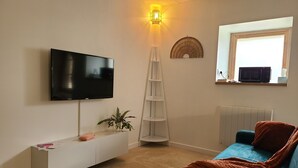 Una sala de estar con un televisor de pantalla plana montado en la pared, un mueble de entretenimiento blanco con una planta, un sofá azul con cojines naranjas y una escalera apoyada contra la pared.