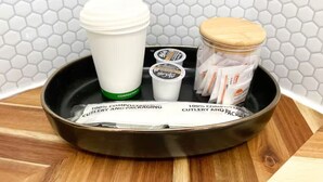 Una bandeja negra con tapa blanca, una taza blanca, un tarro con tapa de madera y algunos productos envasados sobre una superficie de madera frente a una pared de azulejos.