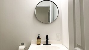 A bathroom with a round mirror, a sink, a hand soap dispenser, and a towel dispenser. - Boutique Hotel - Suite #4 Single (Thunder Bay)