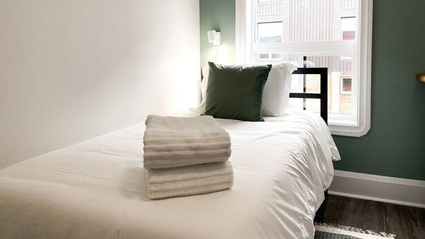 A neatly made bed with a folded white towel, a green pillow, and a white pillow in a room with a window and a view of buildings outside. - Boutique Hotel - Suite #4 Single (Thunder Bay)