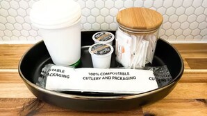 Una bandeja negra con varios objetos, incluyendo una taza blanca, un tarro con tapa de madera y algunos materiales de embalaje con la etiqueta "100% cubiertos y embalaje compostables".