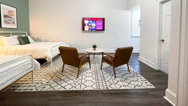 Un dormitorio con una cama, dos sillas, una mesita y un televisor montado en la pared.