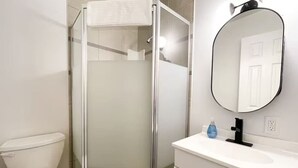 Une salle de bain avec une douche à parois de verre, des toilettes blanches, un lavabo blanc avec un robinet noir et un miroir rond au mur.