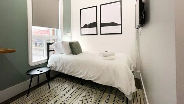 Quarto com uma cama, uma mesinha, uma cadeira, uma janela e duas imagens emolduradas na parede.