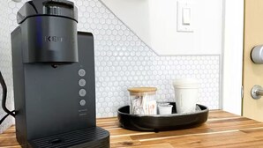 A Keurig coffee maker on a wooden countertop with a hexagonal tile backsplash and a door in the background.