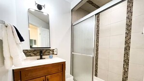 A bathroom with a wooden vanity, a white sink, a mirror, and a glass shower enclosure with mosaic tiles. - Boutique Hotel - #10 Two Double (Thunder Bay)