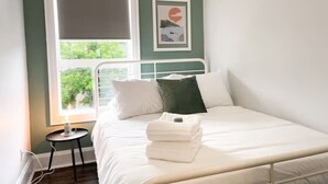A neatly made bed with pillows and a folded towel, a small bedside table with a lamp, and a window with a view of greenery. - Boutique Hotel - #10 Two Double (Thunder Bay)