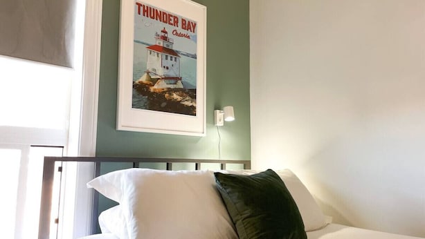 Ein ordentlich bezogenes Bett mit weißen und grünen Kissen in einem Zimmer, an der Wand hängt ein Bild von einem Leuchtturm.