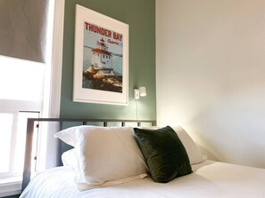 Ein ordentlich bezogenes Bett mit weißen und grünen Kissen in einem Zimmer, an der Wand hängt ein Bild von einem Leuchtturm.