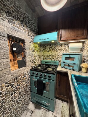 A kitchen with a stone wall, a vintage stove, wooden utensils, and a blue cabinet. - Beautiful Yo Mama’s GuestHouse (Wrightwood)