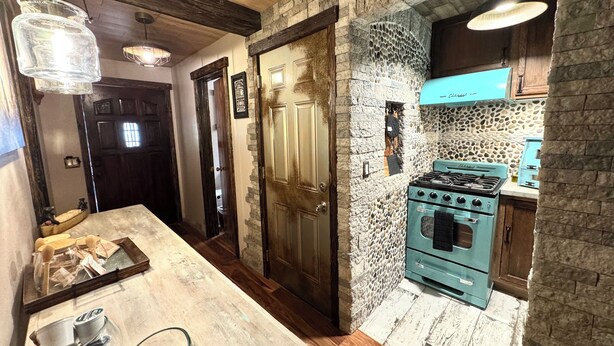 Une cuisine avec un mur en pierre, un poêle d’époque et des poutres en bois au plafond.