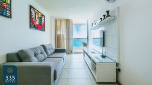 Ein modernes Wohnzimmer mit grauem Sofa, einem Flachbildfernseher und Meerblick durch ein großes Fenster.