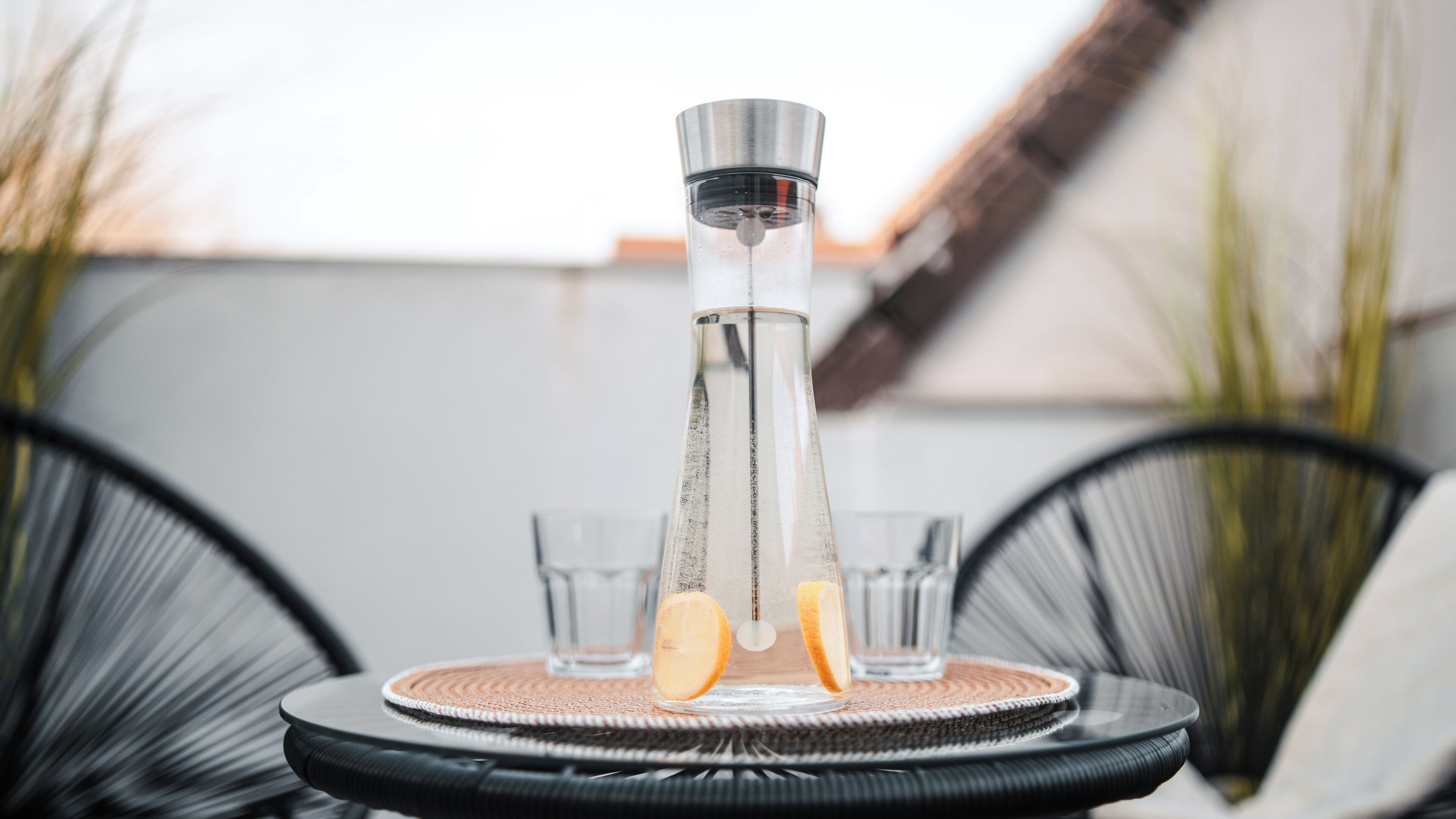 A clear glass bottle with a metal cap, two glasses, and a woven coaster on a round table.