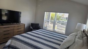 Um quarto com cama, cômoda, televisão, cadeira, janela e abajur.