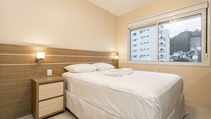 A neatly made bed with a headboard, a bedside table with drawers, and two wall-mounted lamps. - 50" Smart TV | 500 meters from UFSC #TRINA03D (Florianópolis)
