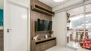 A modern living room with a flat-screen TV mounted on a wooden wall unit, a sliding glass door leading to a balcony, and a white door. - 50" Smart TV | 500 meters from UFSC #TRINA03D (Florianópolis)