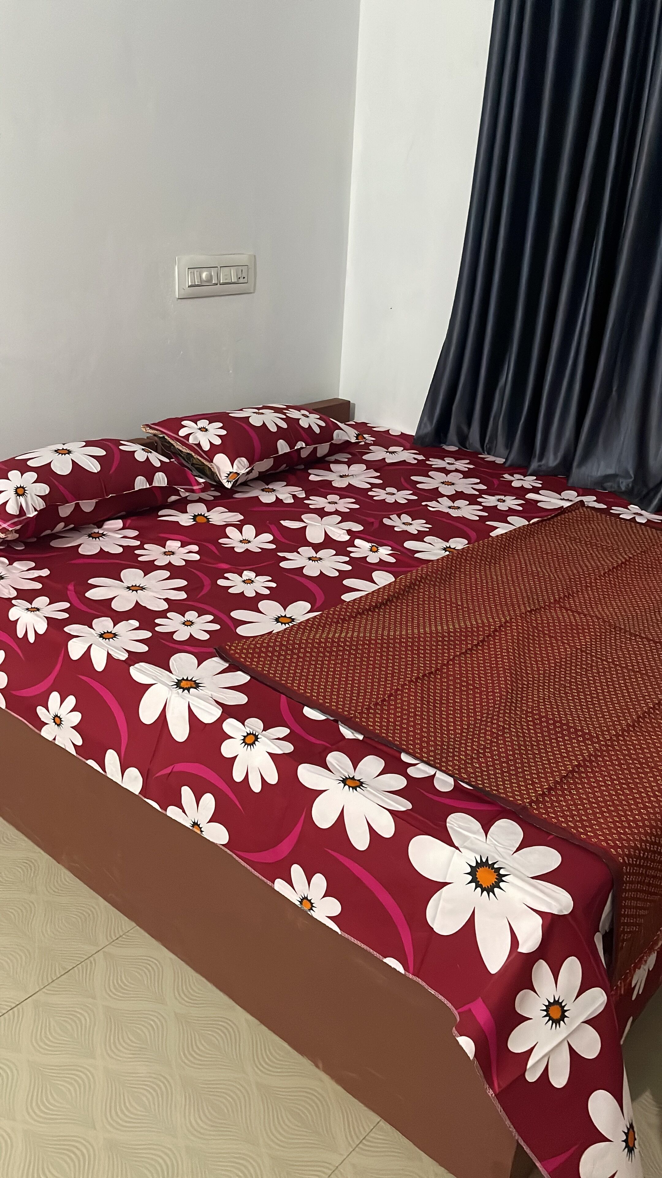 Un lit avec une couvre-lit à motifs floraux, une prise électrique sur le mur et un rideau foncé.