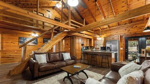 A spacious cabin interior with a wooden staircase, a living area with a brown leather sofa, a coffee table, and a kitchen area with a refrigerator and bar stools.