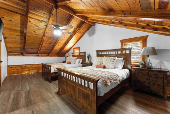 A bedroom with a sloped ceiling, wooden flooring, two beds, a nightstand, and a window with blinds.
