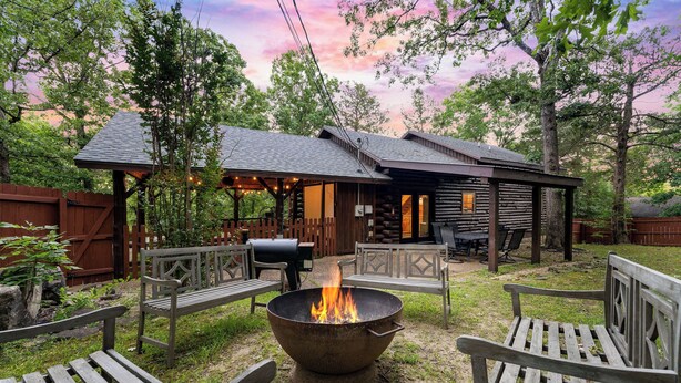 A rustic cabin with a fire pit in the center, surrounded by wooden benches and a covered patio area.