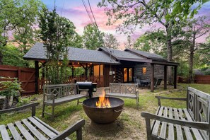 A rustic cabin with a fire pit in the center, surrounded by wooden benches and a covered patio area.