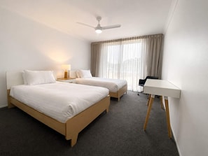 A hotel room with two beds, a desk, a ceiling fan, and a window with curtains.